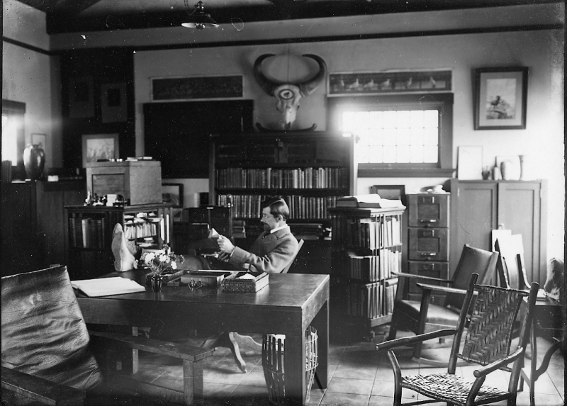 an old black-and-white photograph of an office with furnishings from the turn of the century. A man with glasses and a small pointed beard sits reading behind a desk with his feet up. Books are prominent, as is the skull of a horned animal mounted on the wall. Sunlight streams in through a small window.