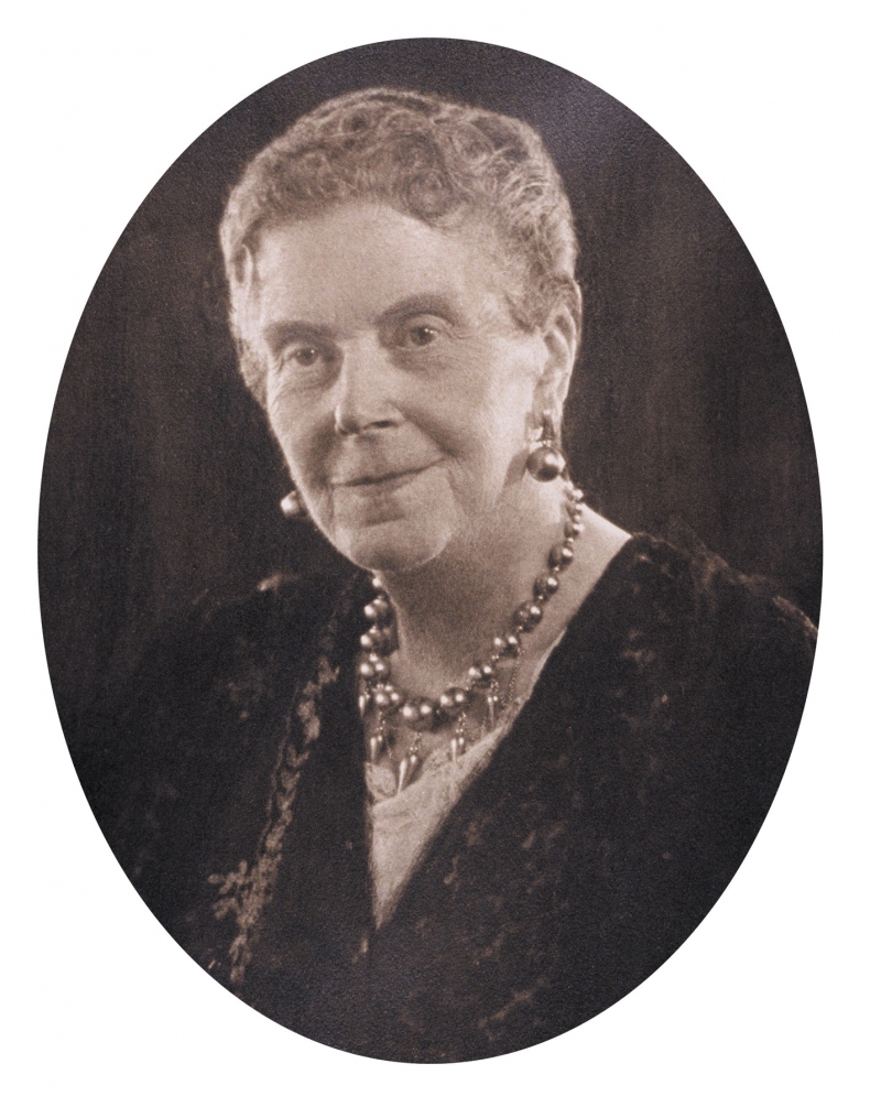 A sepia-toned old photograph of a smiling elderly woman with short gray hair wearing fancy jewelry.