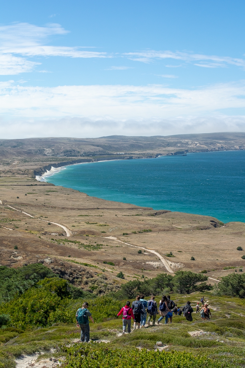 An inspiring view looking out over the north side of Santa Rosa Island. You're looking down on a beautiful bay full of deep blue water. Puffy clouds float in the sky overhead. A dirt road stretches out in the distance over flat bluffs by the sea. In the foreground, plants grow low to the ground, the landscape looks windy. A line of people, mostly teens, are hiking on a trail.