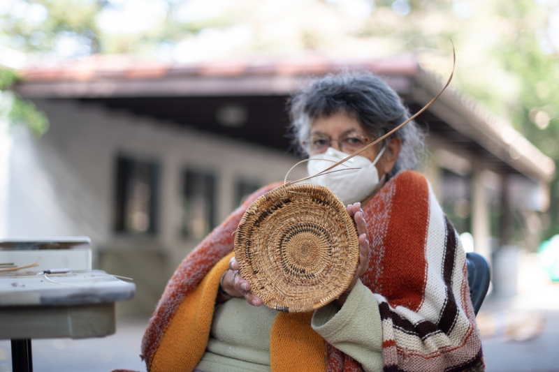 A woman with grey hair and an arched eyebrow holds up a small basket. The stitches have different textures and there are variations in the colors. Strands coming off it show the basket is unfinished. She wears a mask because of the 2020 pandemic.
