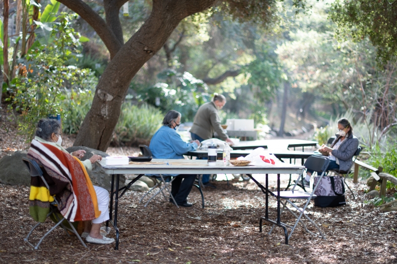 Four women sit apart from each other at tables spread out in the garden. They wear masks because of the 2020 pandemic. Each is busy with something in her hands: basketweaving