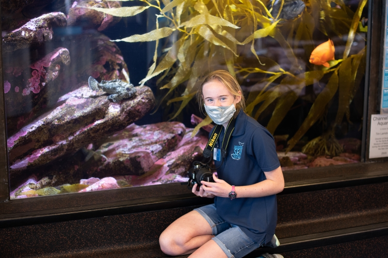Lucy is holding a camera and kneeling in front of an aquarium tank. She wears a badge and a shirt with the Sea Center logo on it. Behind her in the tank are fish, giant kelp, and crusty pink algae