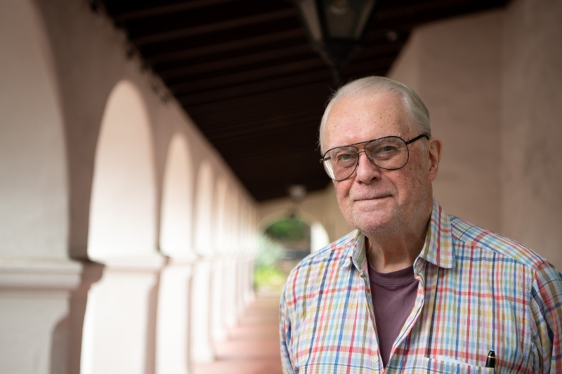 A smiling older man wearing glasses and a Madras plaid shirt of many colors. He stands in front of a row of columns at the Museum.