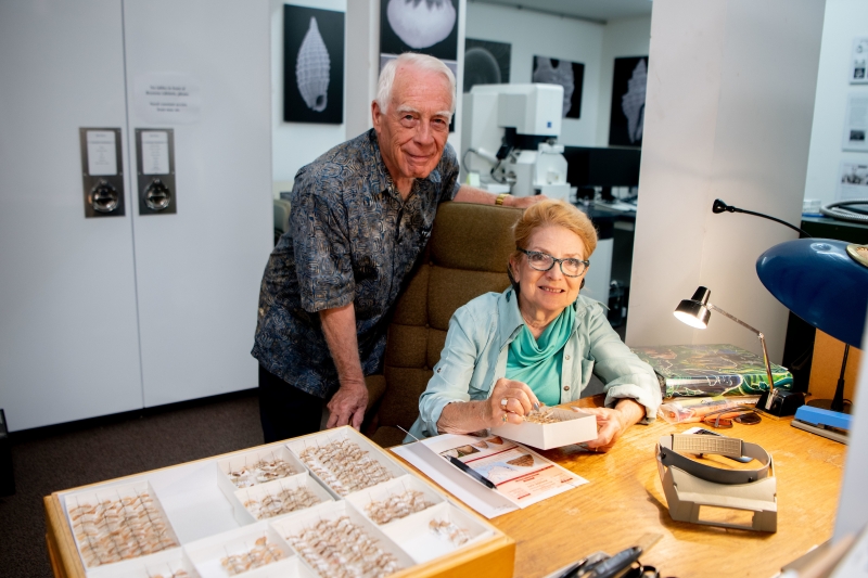 A smiling older couple in a scientific lab setting with a drawer of pinned moths