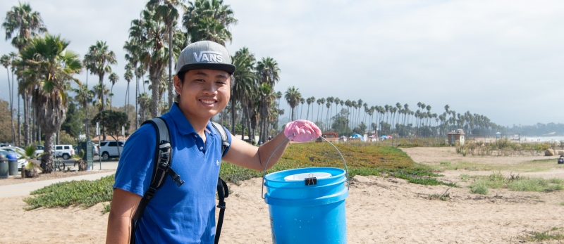 A smiling young man holds up a blue bucket of trash on the beach