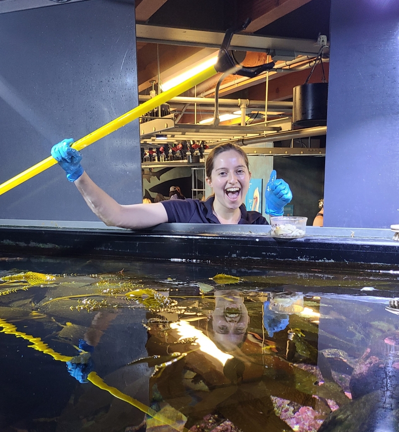 A smiling young woman gives the thumbs up as she holds a long grabbing tool for use feeding fish over an open aquarium