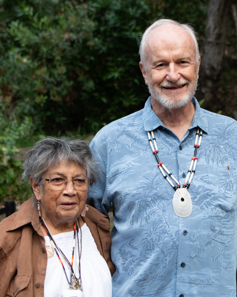 A petite older woman standing with a tall man, both wearing beaded necklaces