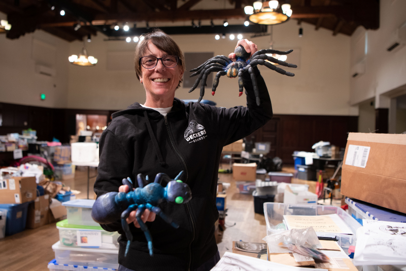 A smiling woman holds up giant spider demonstration models in a large auditorium filled with bins and boxes of supplies.