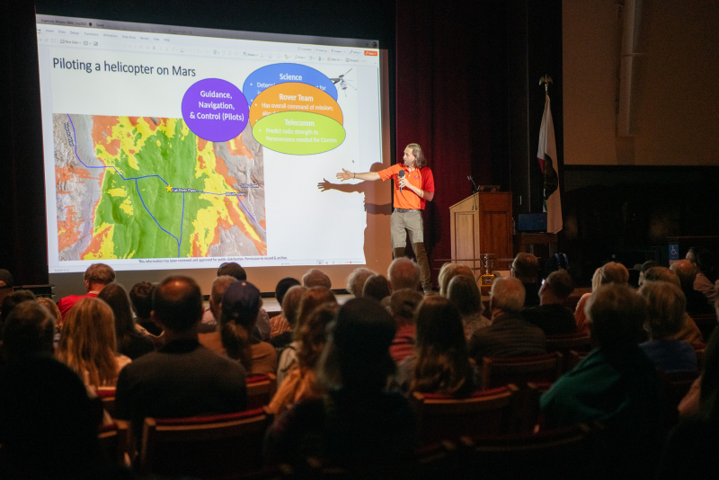 A man in a bright orange shirt presents a slideshow to an audience in an auditorium