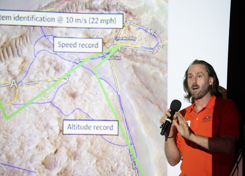 A closeup of a man in a bright orange shirt presenting a slide showing a map of Mars with places where speed and altitude records were set