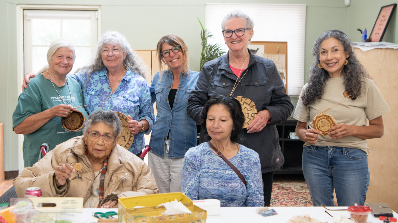 Seven women in a brightly-lit room holding the baskets they are working on