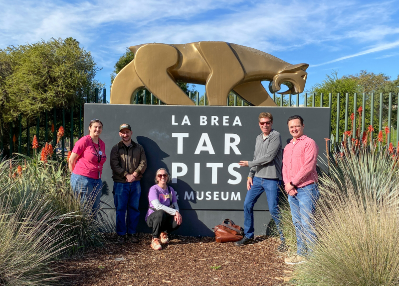 Five smiling people standing around a golden statue of a saber-toothed cat at a sign that reads La Brea Tar Pits & Museum