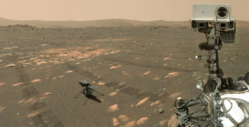 An image from the surface of Mars with the rover and helicopter amid a dusty landscape and rover tracks in the foreground and bare slopes in the distance