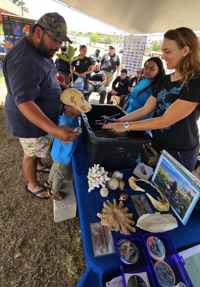 Smiling young woman at an outreach table shows a live sea star to a man and a boy