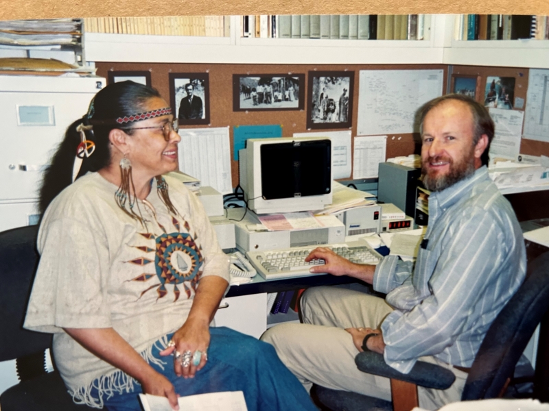 A smiling woman wearing beautiful Native American jewelry and glasses sitting in an office with a bearded man in front of an early computer. Historic photos and books cover the walls behind them.