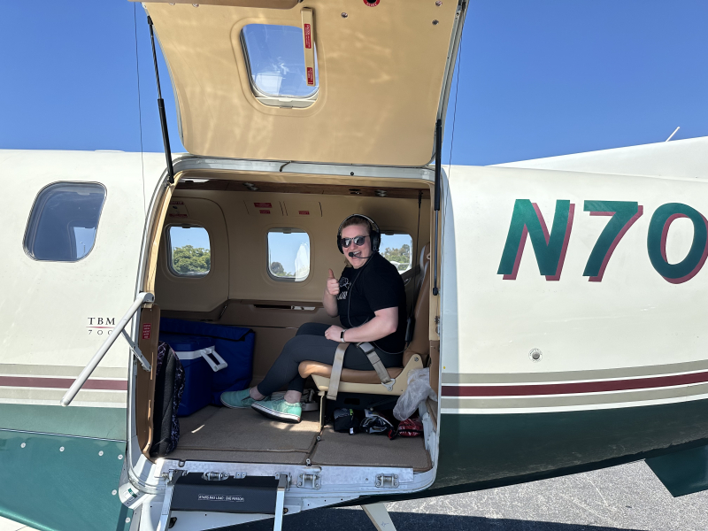 A scientist gives the thumbs up from inside a small aircraft, next to a cooler containing a live endangered snail