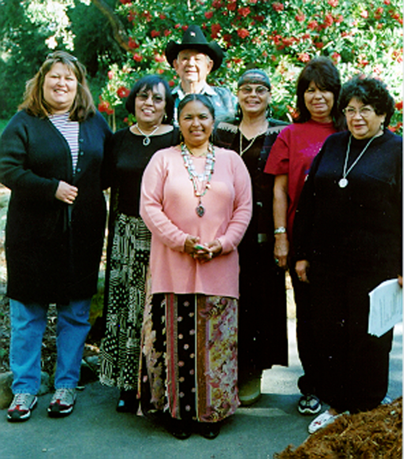 Six smiling women and a smiling man stand together in front of a Toyon plant covered with red berries.