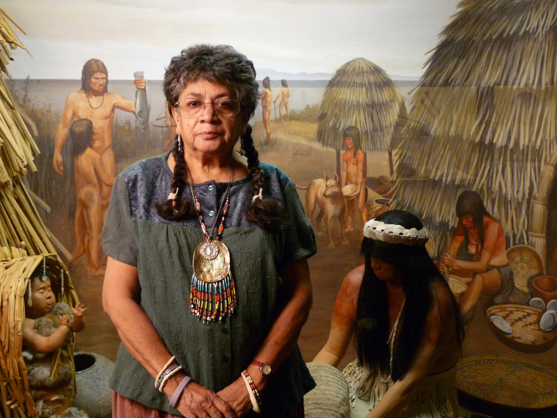 A woman looking confidently into the camera in front of a museum diorama depicting daily life in a Chumash village on the coast. She wears a striking beaded shell pendant and has dark braids.