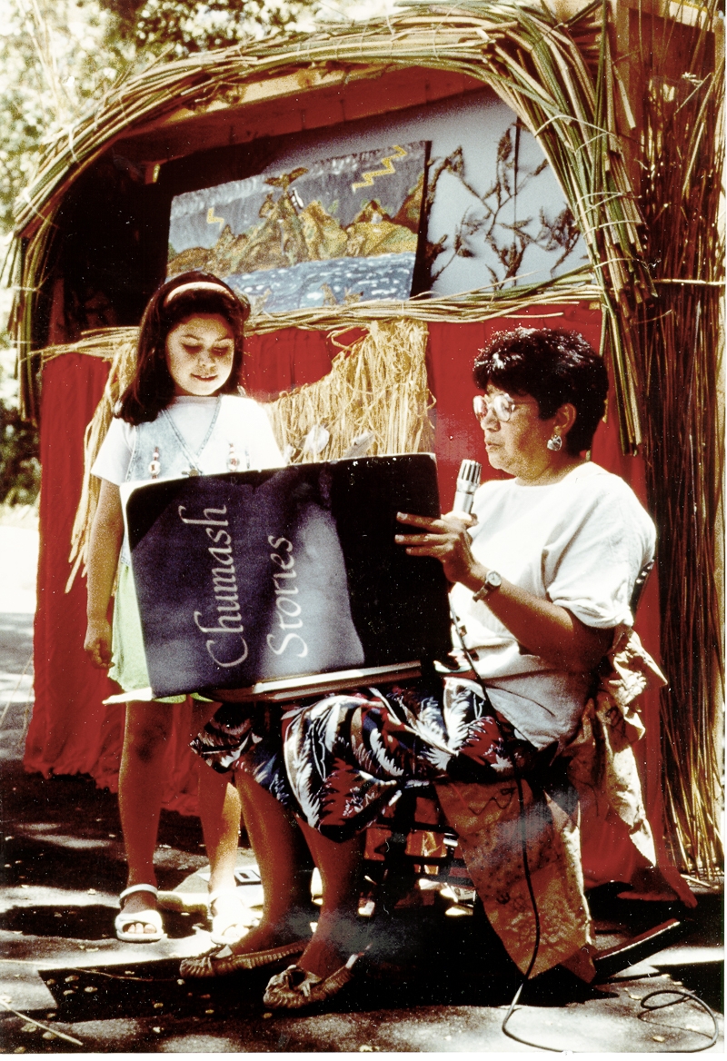 A woman with a microphone reads to a little girl from a book of Chumash stories. They are in front of a small outdoor theater, with a background showing a mountain, lightning bolts, and water. Tule rushes form the proscenium arch.
