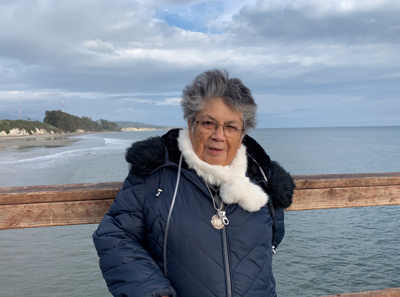 A smiling elder woman bundled up warmly on a cold winter day, standing on a pier overlooking the ocean and a partly cloudy view of the coast.