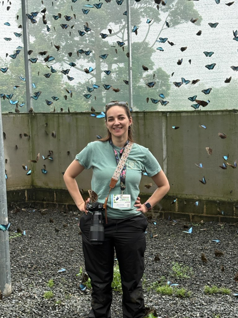 The author smiles as she stands inside a large, mesh-enclosed area absolutely full of large blue butterflies.