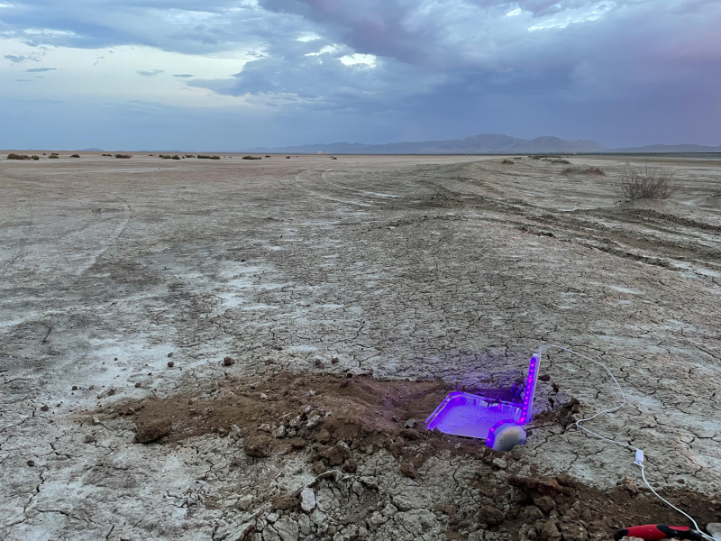 a light trap glows purple at the edge of a barren-looking flat, with stormy clouds overhead