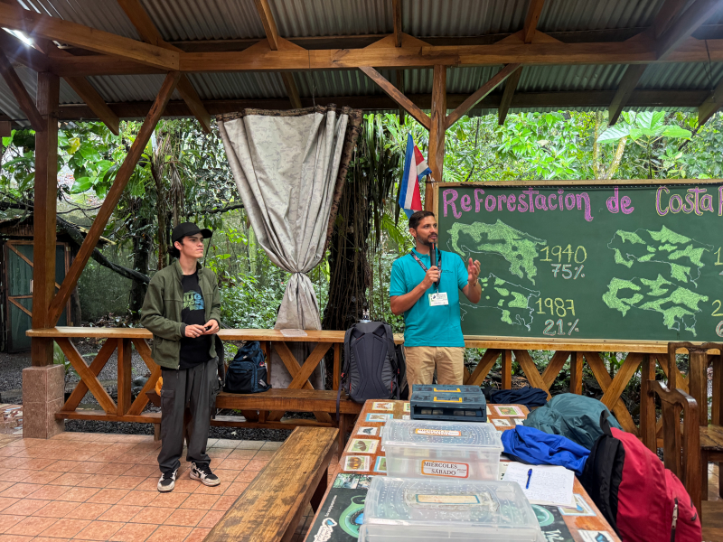 Two men stand by a blackboard with maps showing reforestation over time. They're on a tiled patio with dense vegetation in the background.
