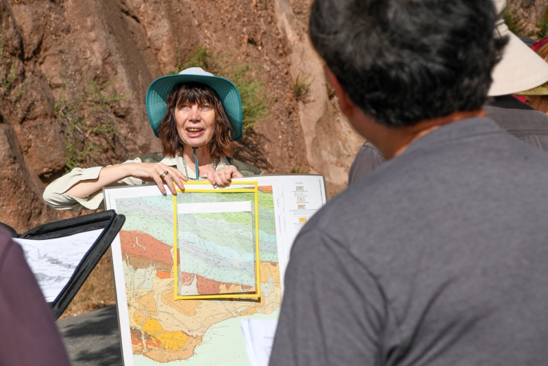 A smiling woman wearing a hat under bright sun holds a colorful map in front of a roadcut, where the rock layers stand out clearly. You're looking at her over the shoulders of students gathered around on the field trip.