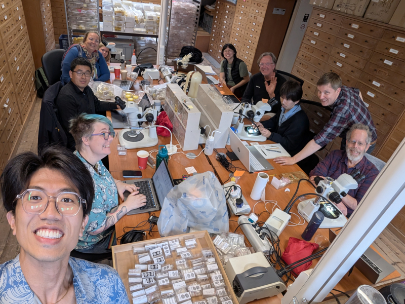 10 smiling scientists clustered around a table full of microscopes and specimens, surrounded by cabinets