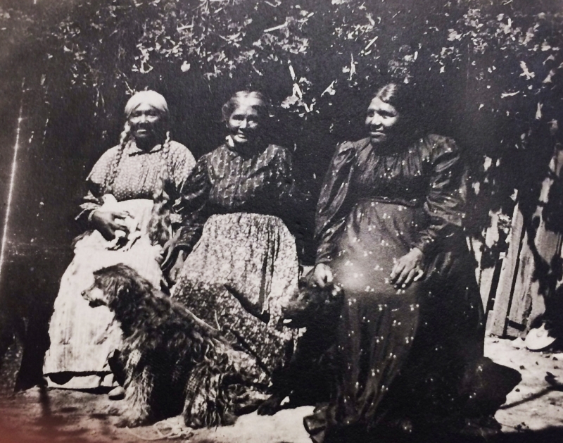 A black and white photo of three seated, smiling women wearing dresses and aprons in an old-fashioned style. A dog sits beside them.