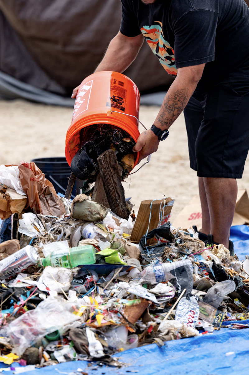 A bucketful of trash being emptied into a big pile of garbage, including plastic wrappers, plastic bottles, and much more.
