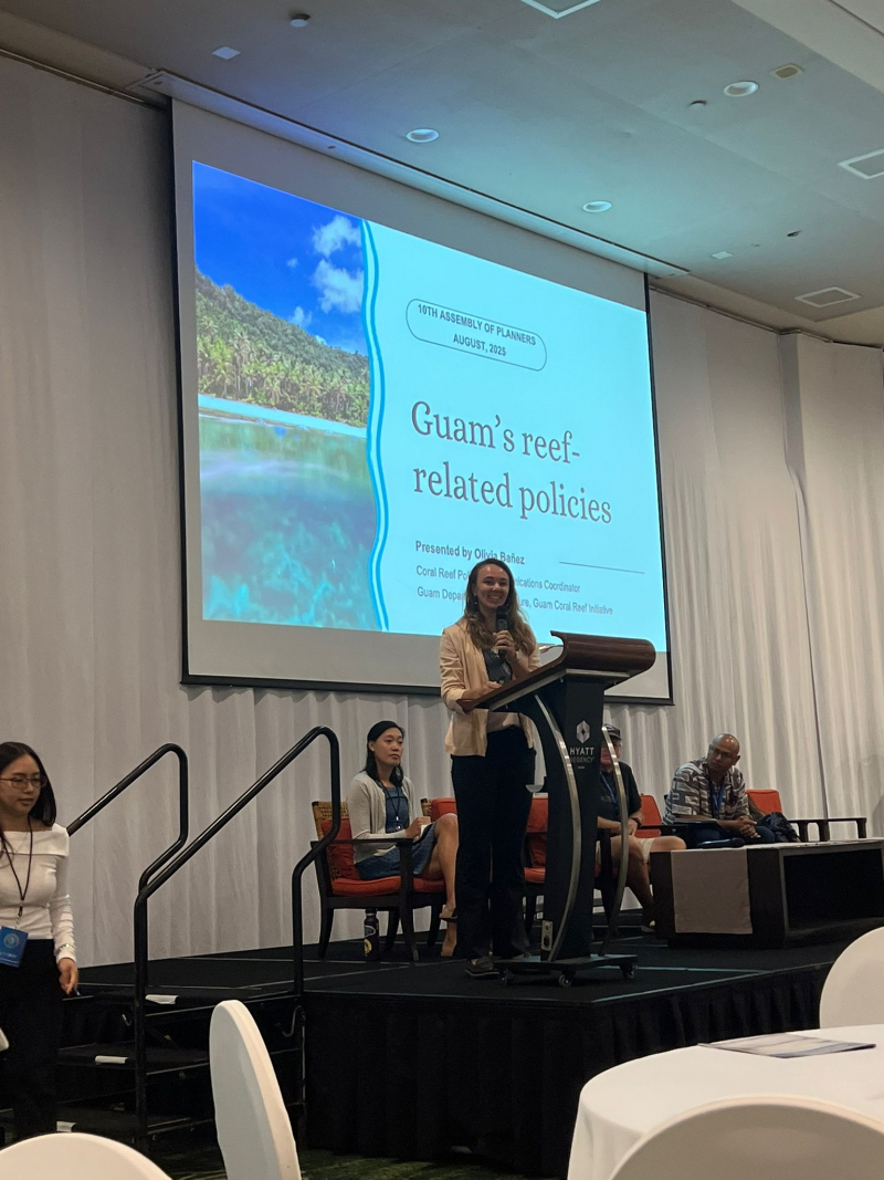 Smiling young woman speaking in front of a slide presentation and panel onstage