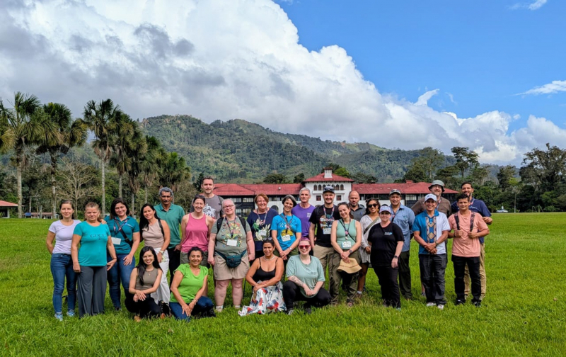 A large group of people in a grassy field with mountains, jungle foliage, and dramatic clouds in the background
