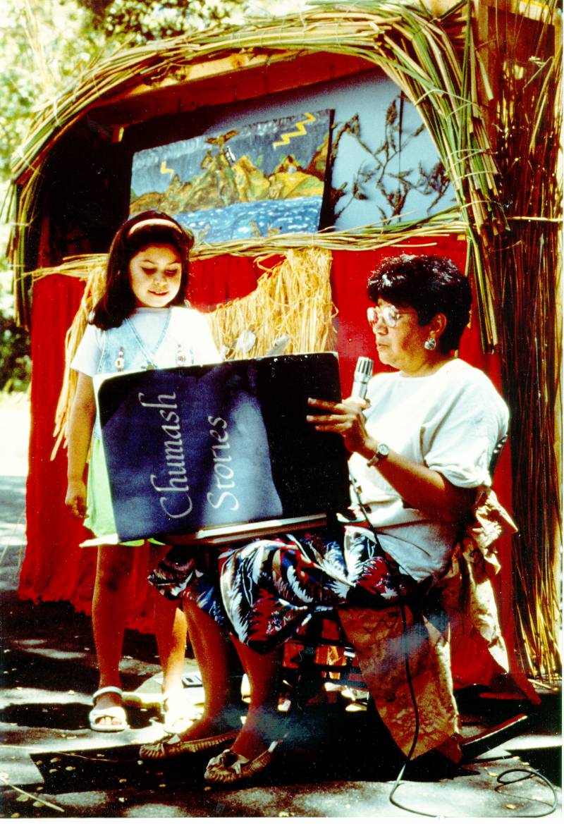 A woman with a microphone reads to a little girl from a large book labeled Chumash Stories, in front of a small theater arch made of tule reeds