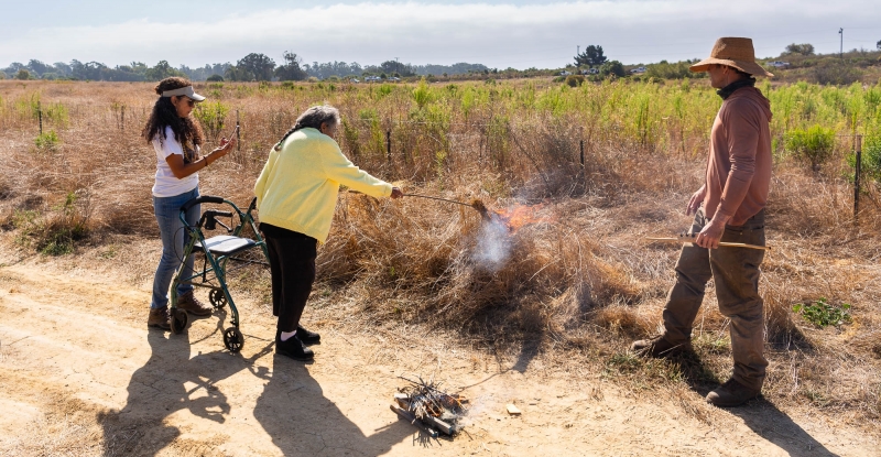 Ernestine Ygnacio-De Soto lights the brush with a firebrand as her niece Samantha Sandoval and Wayne Chapman look on.